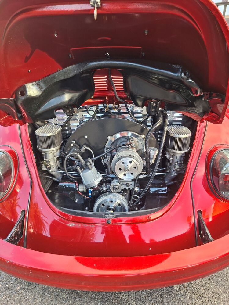 A close-up view of a highly modified and detailed dual-carburetor air-cooled VW Beetle engine bay inside a candy apple red vintage car, showcasing polished chrome pulleys, custom air filters, and clean black shroud components at a car club run.