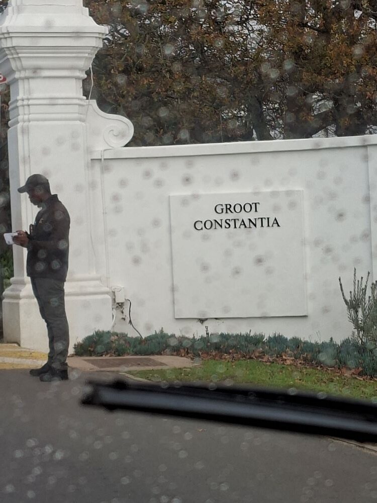 A view through a raindrop-covered car windscreen showing the white entrance wall and pillars of Groot Constantia wine estate, with a security guard standing nearby.