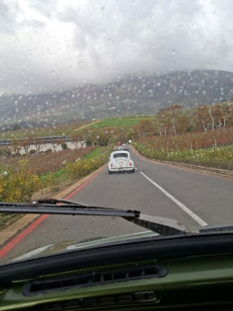 A view through a rain-spotted windscreen with a wiper blade, showing a classic white VW Beetle leading a convoy up a road lined with vineyards toward misty mountains.
