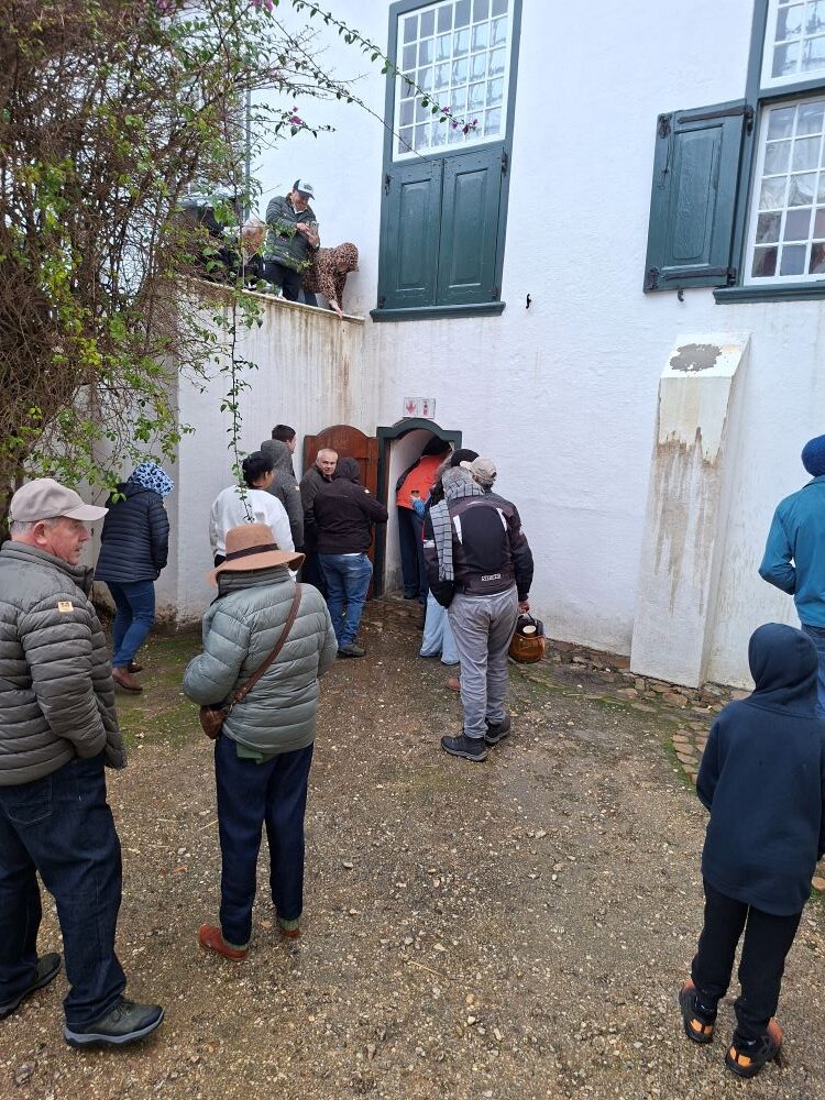 A group of car club members queueing outside a small, low wooden doorway leading under a historic white Cape Dutch building during an estate tour.