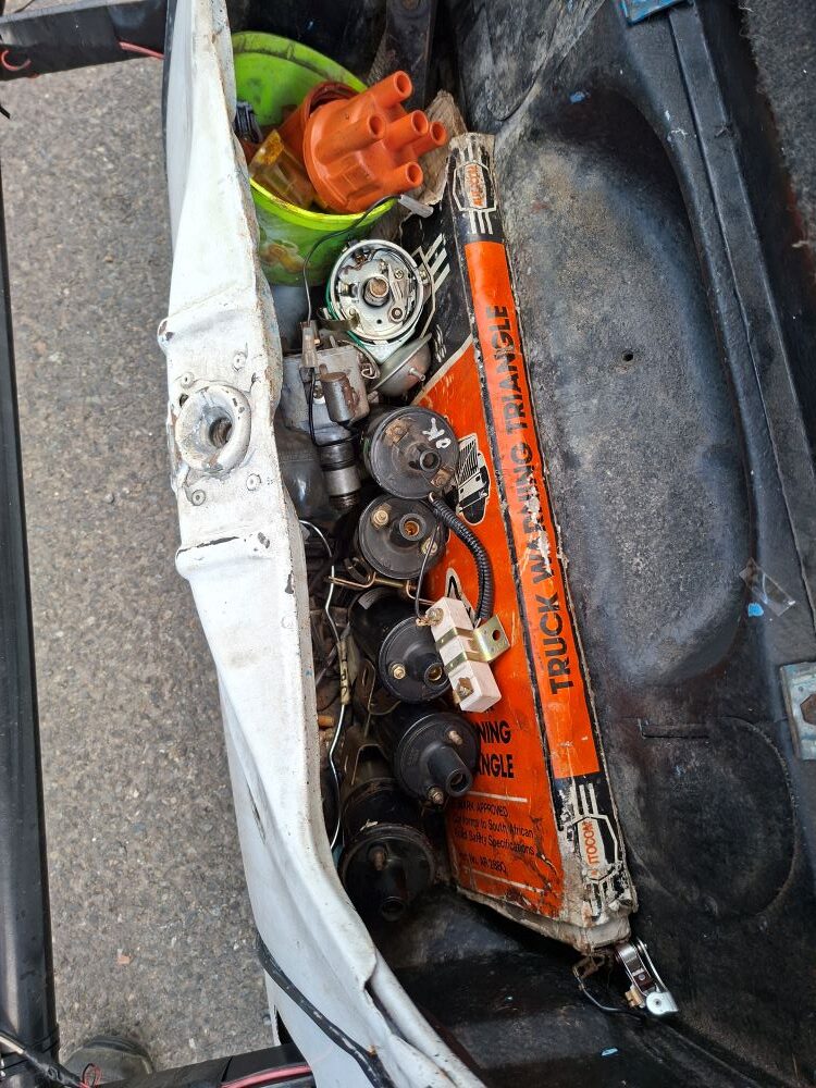 A top-down view of a white vintage classic VW Beetle trunk packed with mechanical spares, featuring a stack of about eight black ignition coils, an orange distributor cap, and an orange warning triangle box packed tightly together for roadside emergency repairs during a Western Cape car club run.