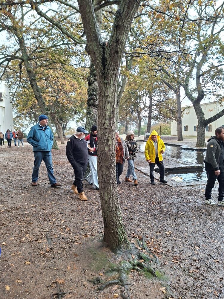 A group of car club members walking along a damp, leaf-strewn path under large autumn trees next to a reflection pool during an outdoor estate tour.