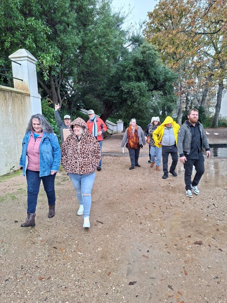 A group of car club members walking together down a wet dirt path under trees next to an estate wall, with one member in the background smiling and making a hand gesture.