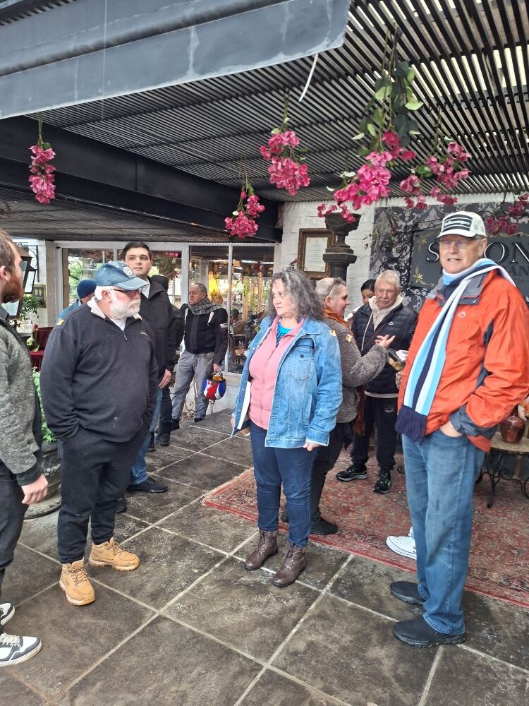 A group of car club members standing on an outdoor covered patio under hanging pink flowers, chatting together outside Simon's Restaurant at an estate.