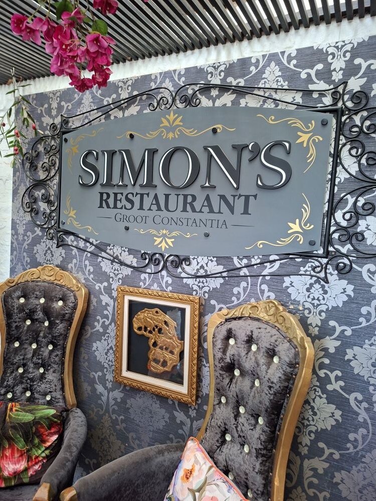 An elegant grey and gold signage for Simon's Restaurant at Groot Constantia mounted on a damask pattern wall above ornate tufted armchairs and a framed golden map of Africa.