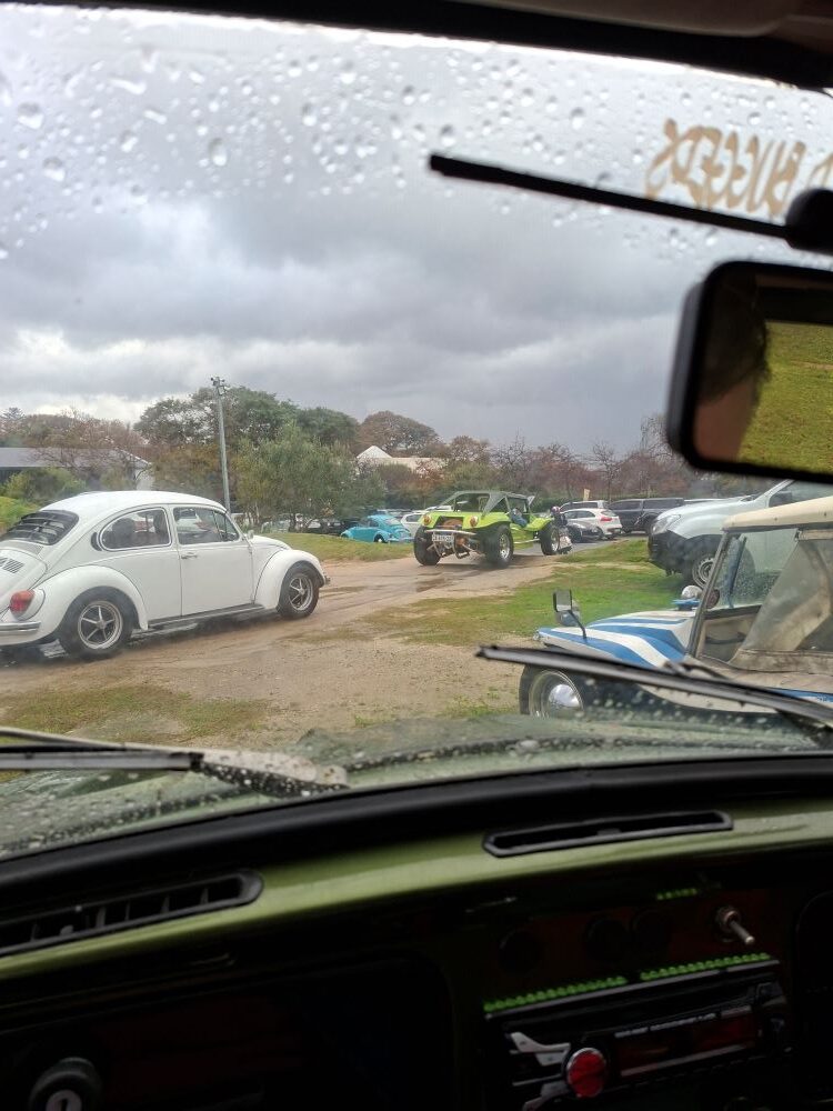 A view from inside a car dashboard looking through a rain-streaked windscreen as a white classic VW Beetle and a lime green beach buggy pull out of a dirt parking lot to start a convoy.