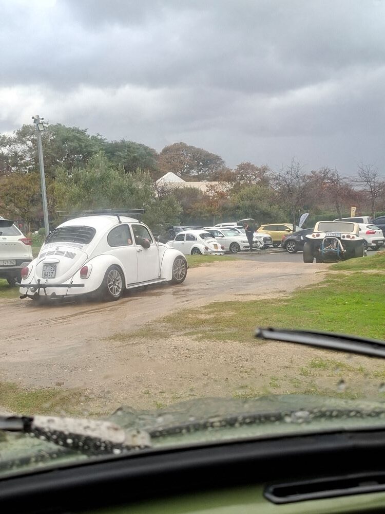 A rear three-quarter view of a white classic VW Beetle with a roof rack and custom alloy wheels driving on a dirt path in a parking area under an overcast sky.