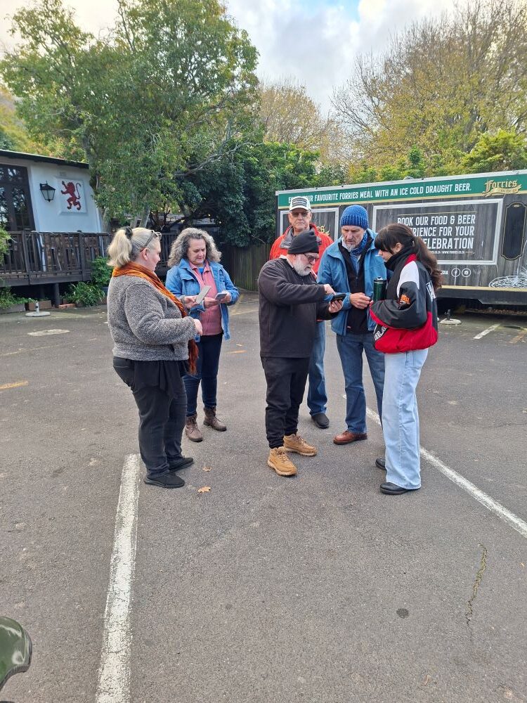 A group of classic car club members standing together in a paved parking lot at a meeting point, huddled around a man showing photos of automotive projects and spare parts on his smartphone screen before an old-school VW cruise.