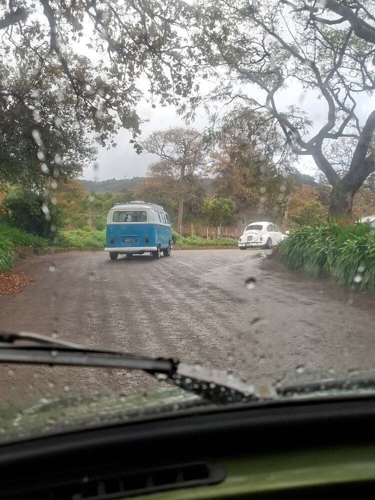 A view through a rain-spotted car windscreen of a classic blue and white VW Kombi and a white Beetle driving along a wet, tree-lined estate road.