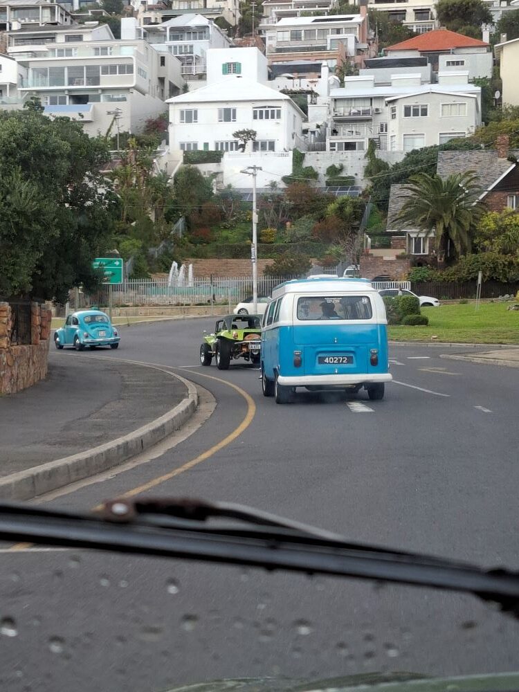 A viewpoint through a rain-flecked car windscreen showing a classic blue and white VW Kombi, a lime green beach buggy, and a light blue Beetle driving along a coastal hillside road in Simon's Town.