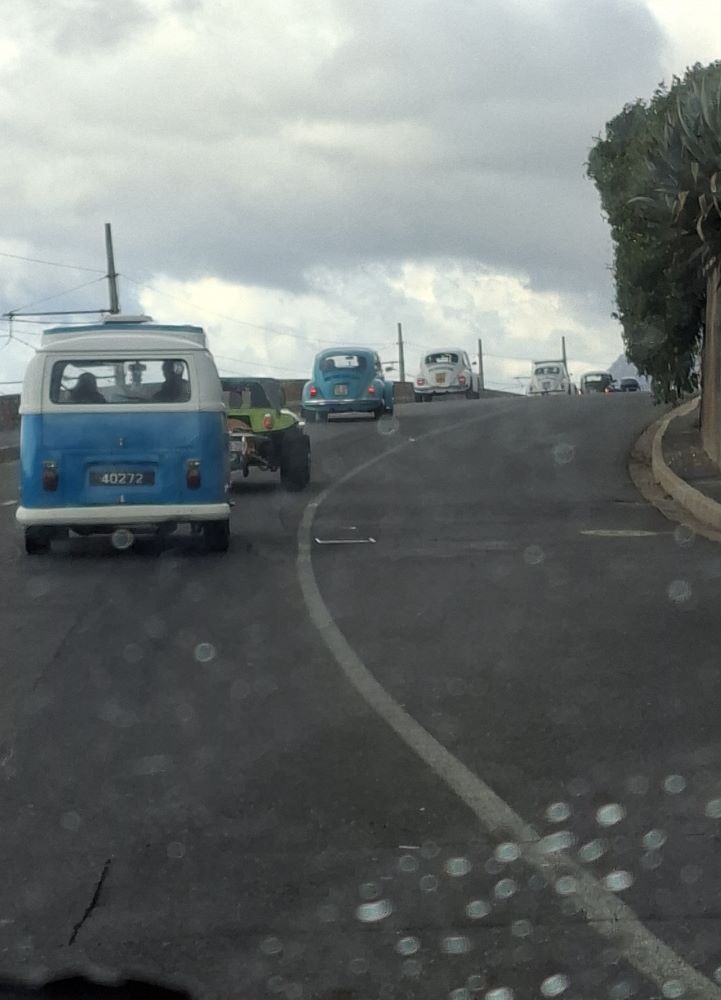 A viewpoint through a rain-dusted car windscreen showing a convoy of classic air-cooled VW vehicles, including a blue and white Kombi, a lime green beach buggy, and several Beetles, driving up a curving coastal road under a cloudy sky.