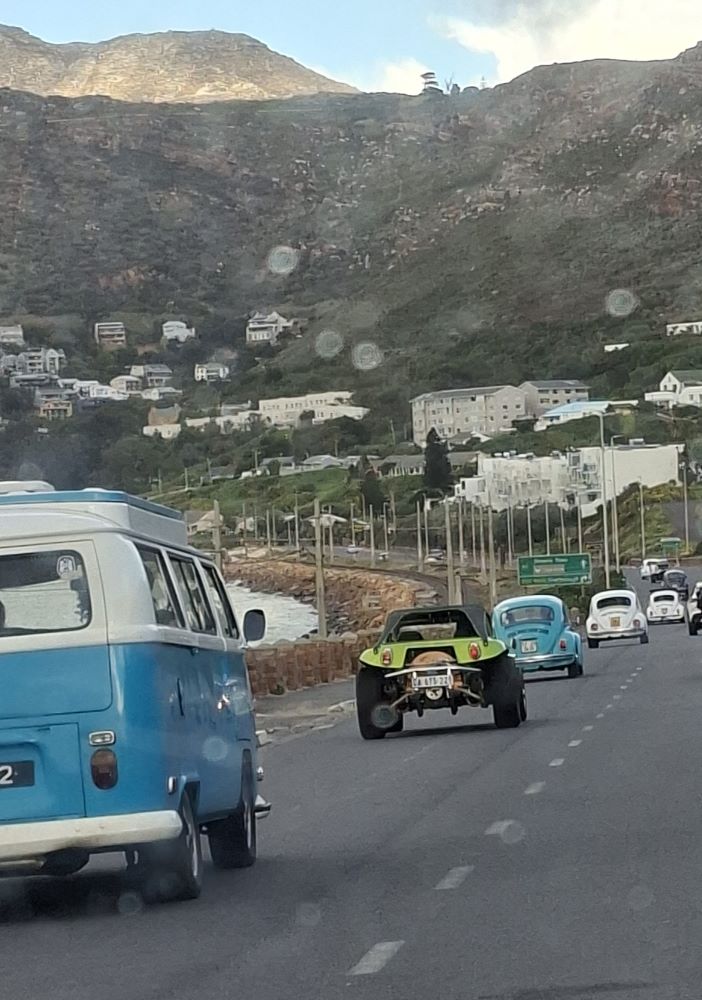 A view from a car windscreen showing a classic blue and white VW Kombi, a lime green beach buggy, and several classic Beetles driving in a convoy along a coastal road next to the ocean in Simon's Town.