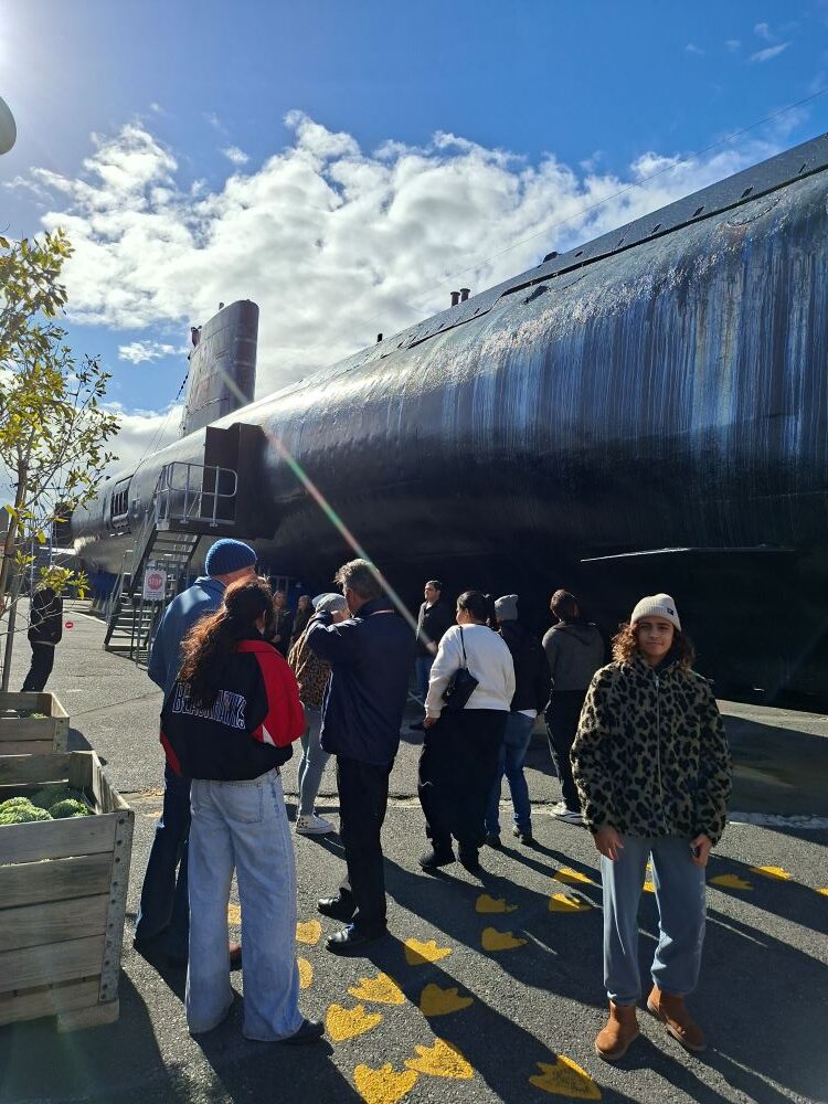 A group of car club members standing on the asphalt outside under a sunny sky, looking up at the massive black hull of a submarine at the naval museum.