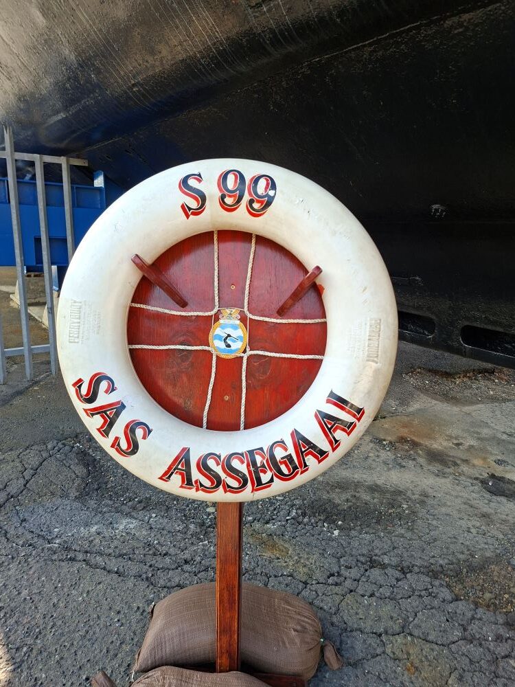 A white and red decorative lifebuoy ring displaying the text S99 SAS Assegaai on a wooden stand, positioned in front of the large black hull of a submarine.
