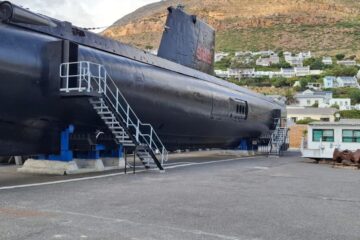 A side profile view of the large black hull and conning tower of the SAS Assegaai submarine resting on supports in a dry dock yard with hills and houses behind it.