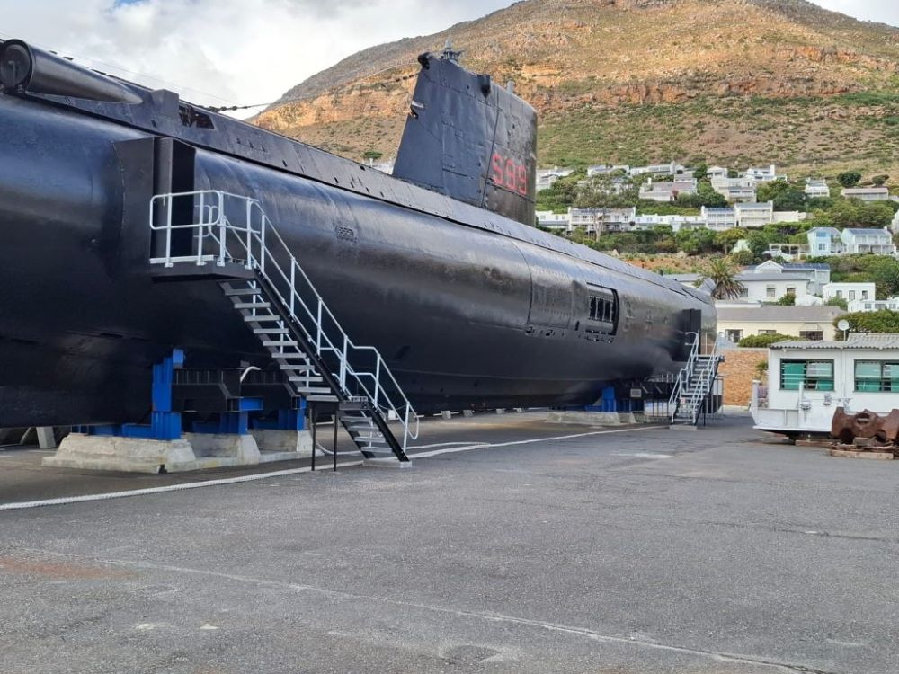 A side profile view of the large black hull and conning tower of the SAS Assegaai submarine resting on supports in a dry dock yard with hills and houses behind it.