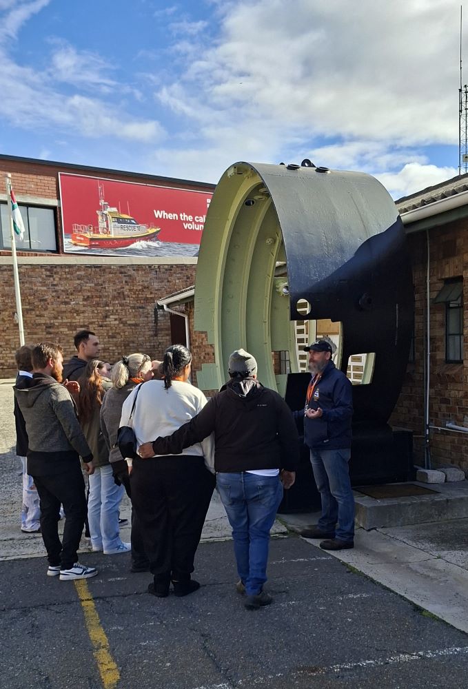 A tour guide speaking to a group of classic car club members standing outside next to a large circular cross-section of a submarine hull at a naval museum.
