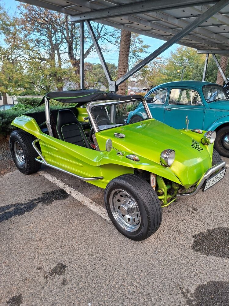 A bright metallic green classic VW beach buggy named Rango with a black soft top roof, chrome wheels, and custom black leather seats, parked next to a blue classic Beetle at a shaded meeting point for a car club event.