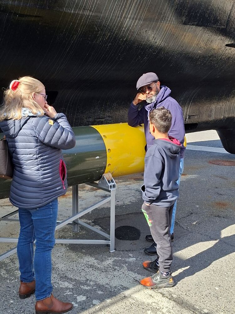 A candid photo of Ras and Clare Abrahams standing with a young boy next to a large green and yellow torpedo display underneath a submarine hull at a naval museum.