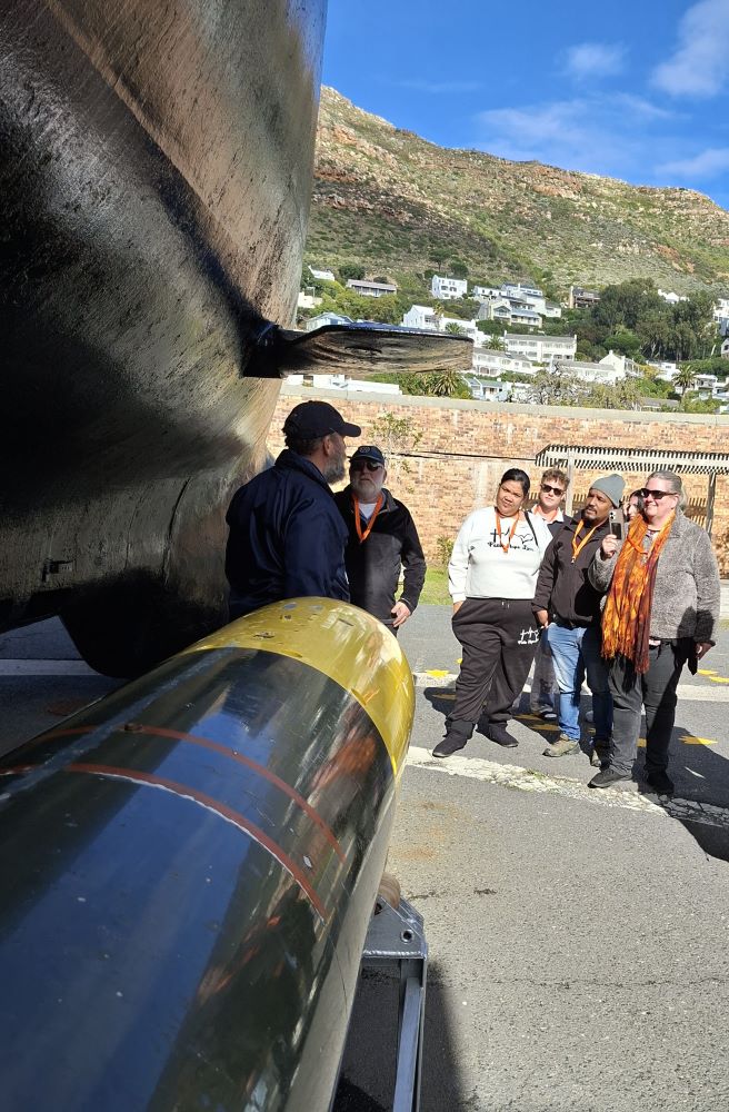 A naval tour guide talking to a group of classic car club members standing outdoors next to a large torpedo display and the massive black hull of a submarine.
