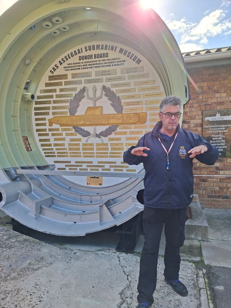 A naval tour guide in a dark jacket gesturing with his hands while standing outdoors in front of a large circular cross-section display of a submarine hull with a donor board inside.