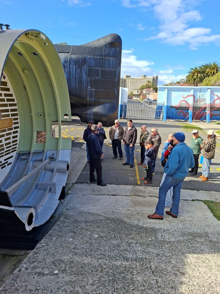 A group of classic car club members gathered in an outdoor courtyard listening to a naval tour guide stand next to a cutaway section of a submarine hull, with the large black stern of a submarine in the background.