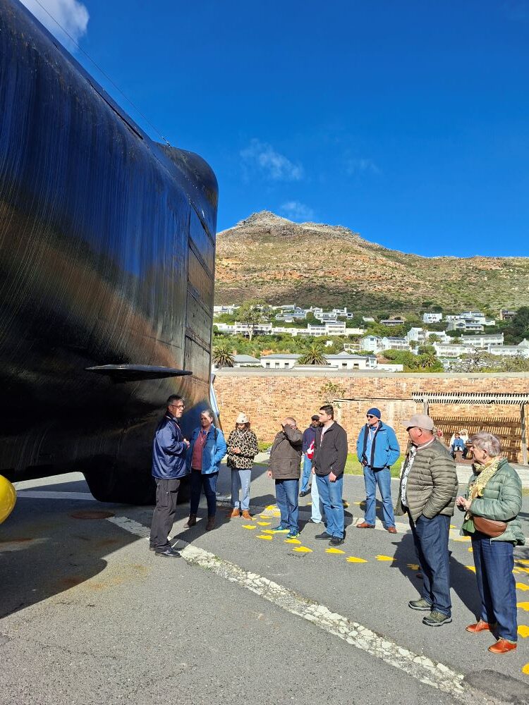 A naval museum tour guide speaking to a group of classic car club members standing on an asphalt courtyard directly next to the massive black stern of a submarine under a bright blue sky.