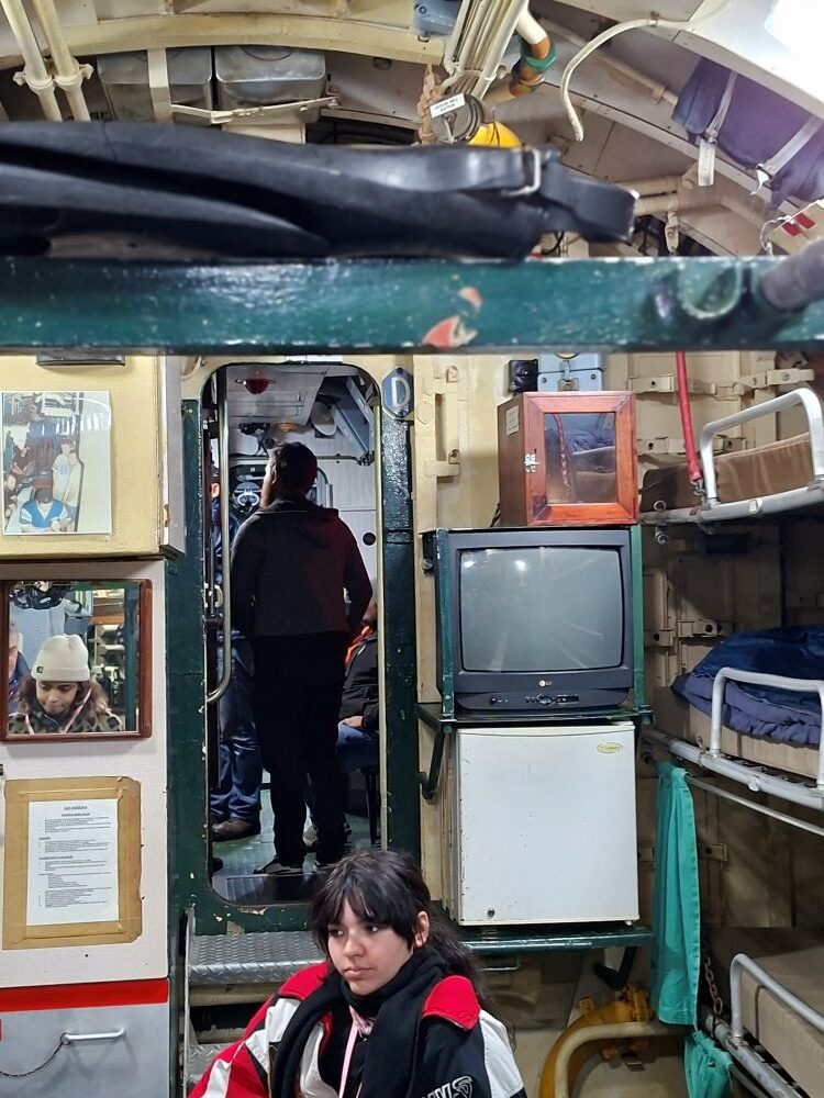 An interior view of the cramped living quarters inside a classic naval submarine, showing stacked bunk beds, an old television set, a small refrigerator, and club members walking through a narrow doorway.