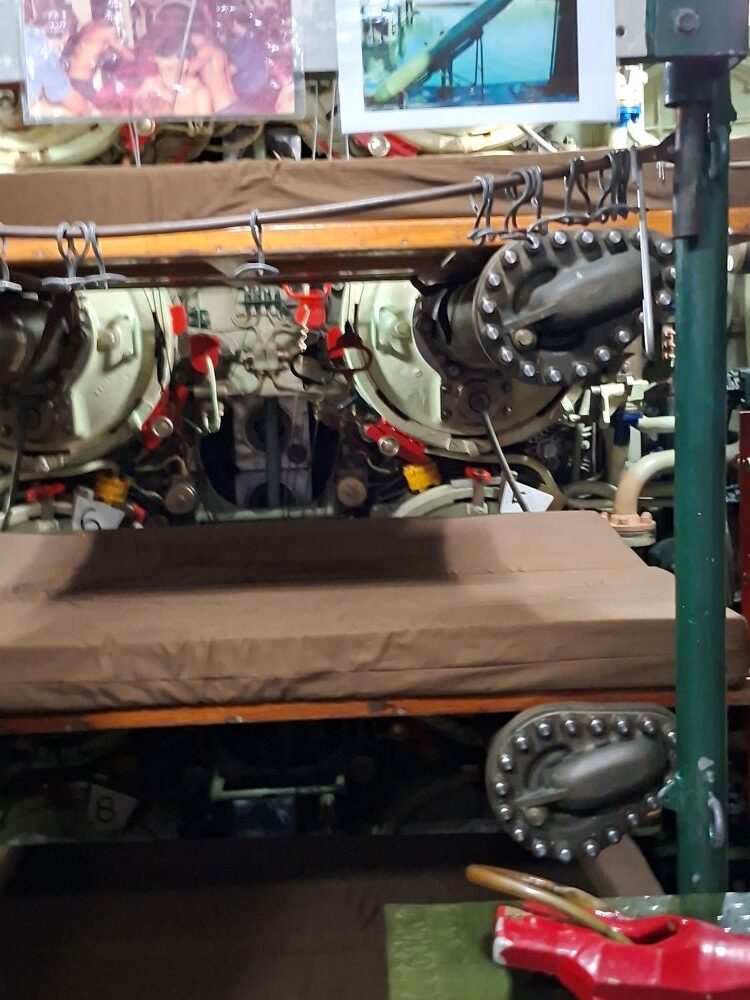 An interior view of the forward torpedo room inside a classic naval submarine, showing stacked crew bunks positioned directly in front of the circular steel torpedo tubes and mechanical launching equipment.