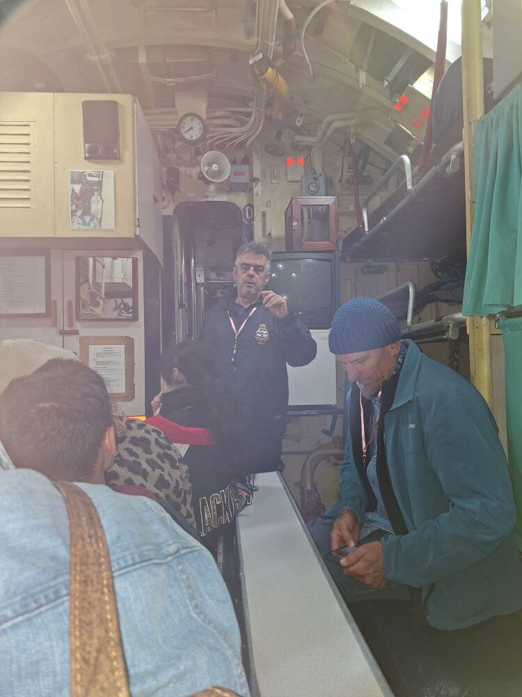 An interior view of the tight crew quarters inside the SAS Assegaai submarine, with a naval tour guide speaking to a group of classic car club members squeezed along a narrow table.