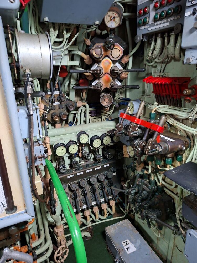 A detailed interior view of a submarine control panel wall covered in a complex array of mechanical switches, circular pressure gauges, heavy levers, and colored wiring bundles.