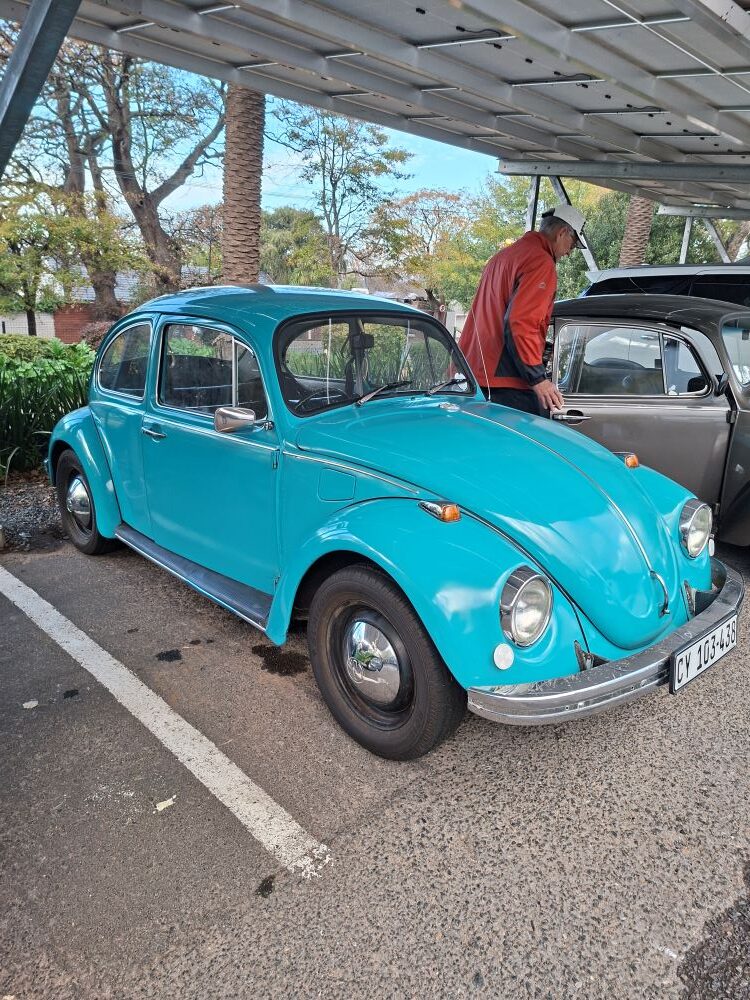 A beautifully restored turquoise blue classic VW Beetle with chrome hubcaps and bumpers, parked in a designated parking bay under a solar panel car port structure next to a grey vintage car at a club meetup point.