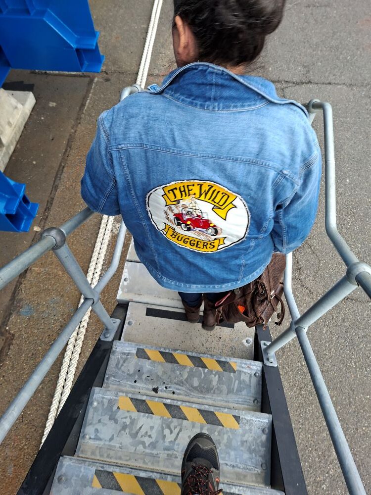 A high-angle view looking down the stairs of a submarine gangway at Mickey wearing a denim jacket with a large embroidered Wild Buggers club patch on the back.