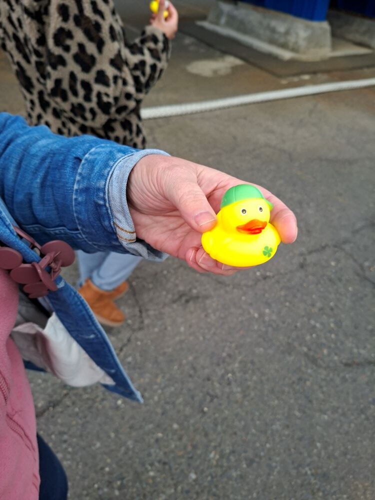 A close-up view of a hand holding a small yellow rubber duck wearing a green cap with a shamrock logo, earned during a submarine tour task.