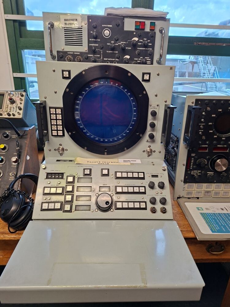 A vintage naval passive tracking sonar console with a large circular blue radar display screen, buttons, and dials on a wooden table at an exhibition center.