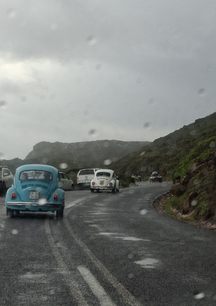 A view through a rain-splattered windshield showing a convoy of classic VW Beetles and beach buggies driving along a wet, winding coastal mountain pass under overcast skies.