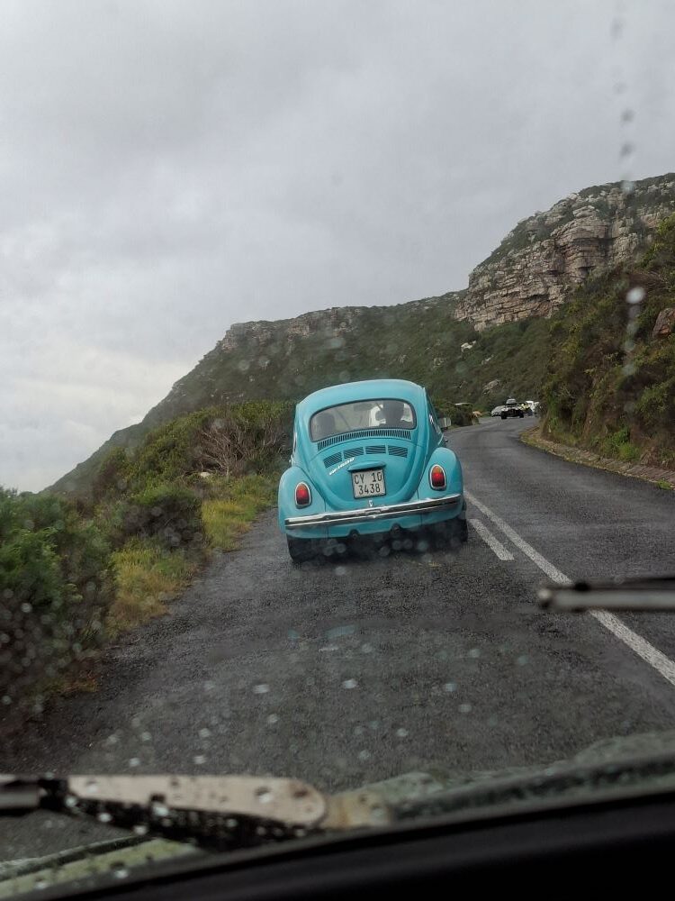 A first-person perspective through a wet windshield showing Heinrich Thiart’s turquoise blue classic 1600 VW Beetle driving along a winding coastal mountain pass in the rain.