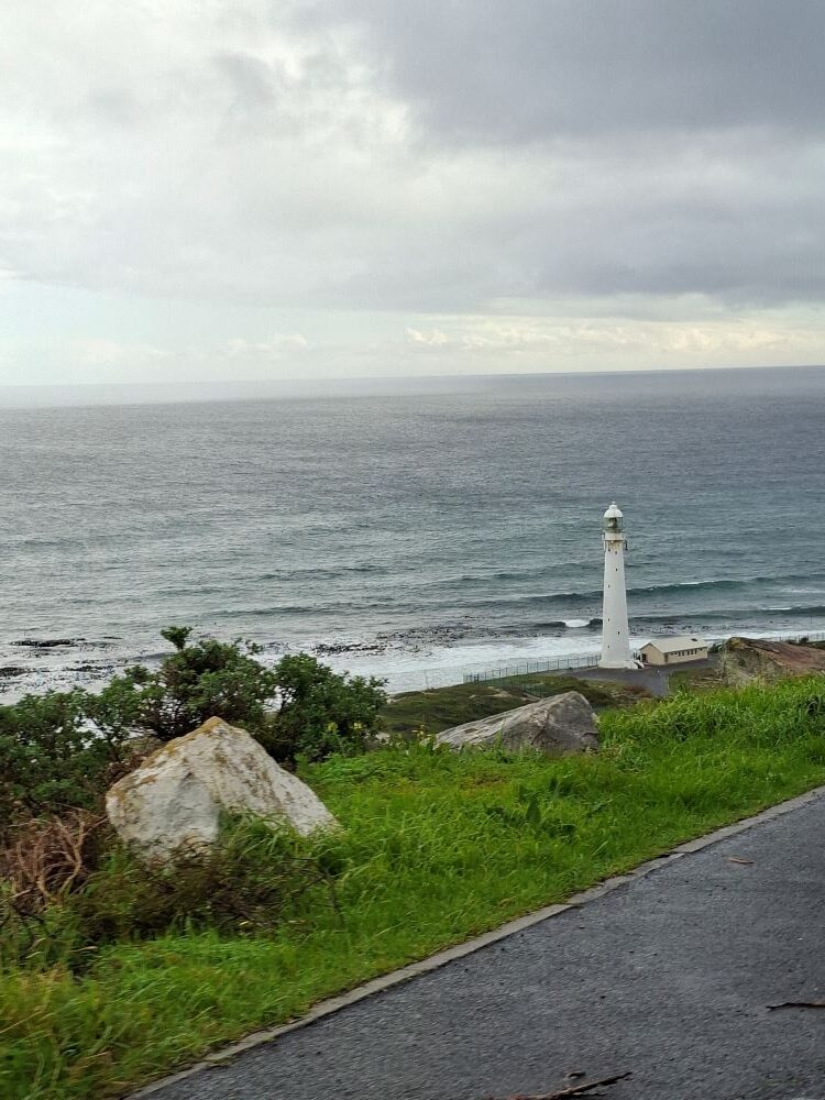 A scenic view looking down from a coastal road at the white Slangkop Lighthouse standing on the rocky shoreline of the Atlantic Ocean under a moody, overcast sky.