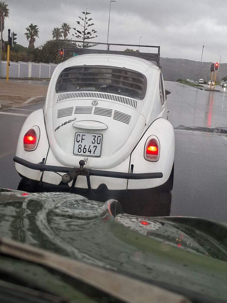 A first-person view from a trailing vehicle looking at a classic white VW 1600 Beetle with a roof rack and tow bar stopped at a wet intersection in the rain.