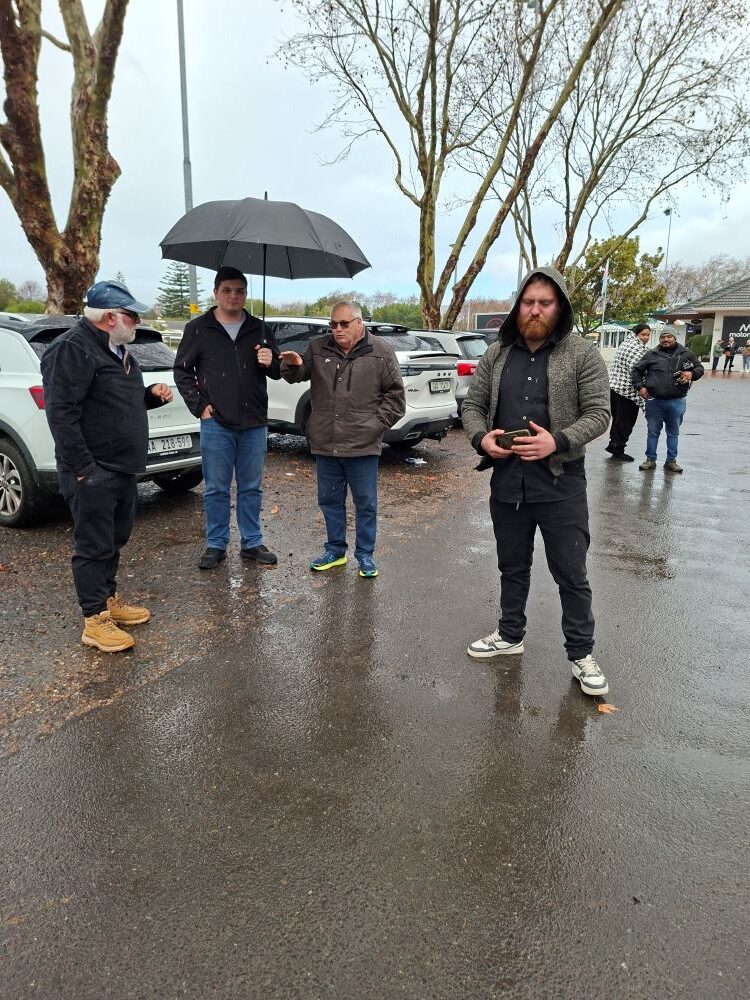 A group of classic car club members standing on a wet asphalt parking lot under a black umbrella, talking and socializing in the rain.