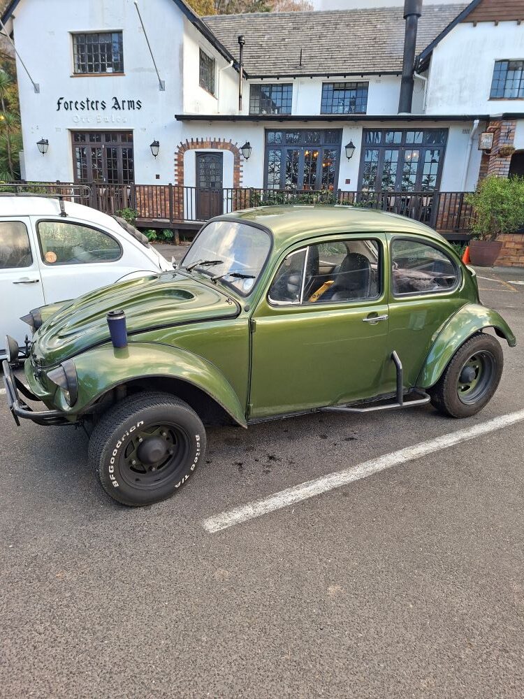 A custom metallic olive green 1974 classic VW Baja Bug named Dark Side with large off-road BFGoodrich tyres, rugged black side bars, and a coffee mug resting on the front wing, parked outside the historic Foresters Arms pub at a car club event assembly point.