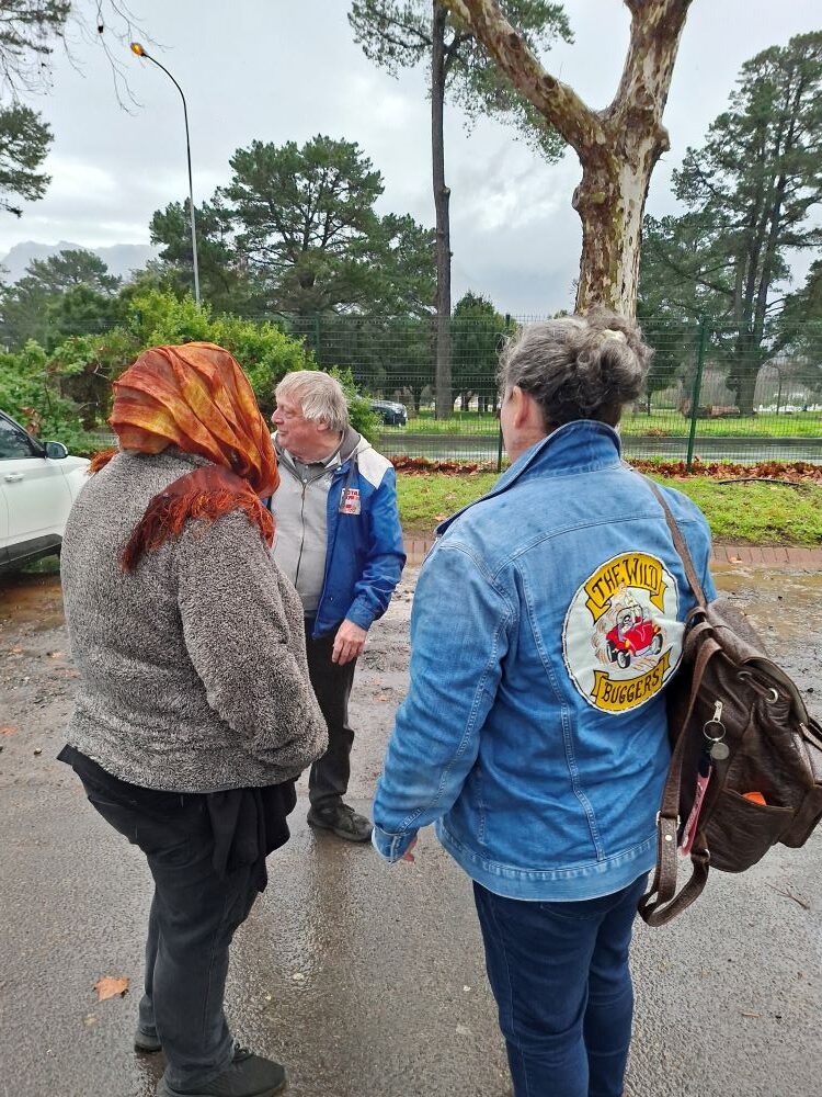 A candid shot of classic car club members, including Mickey in her branded denim jacket, standing on a wet asphalt lot chatting during a rainy day out.