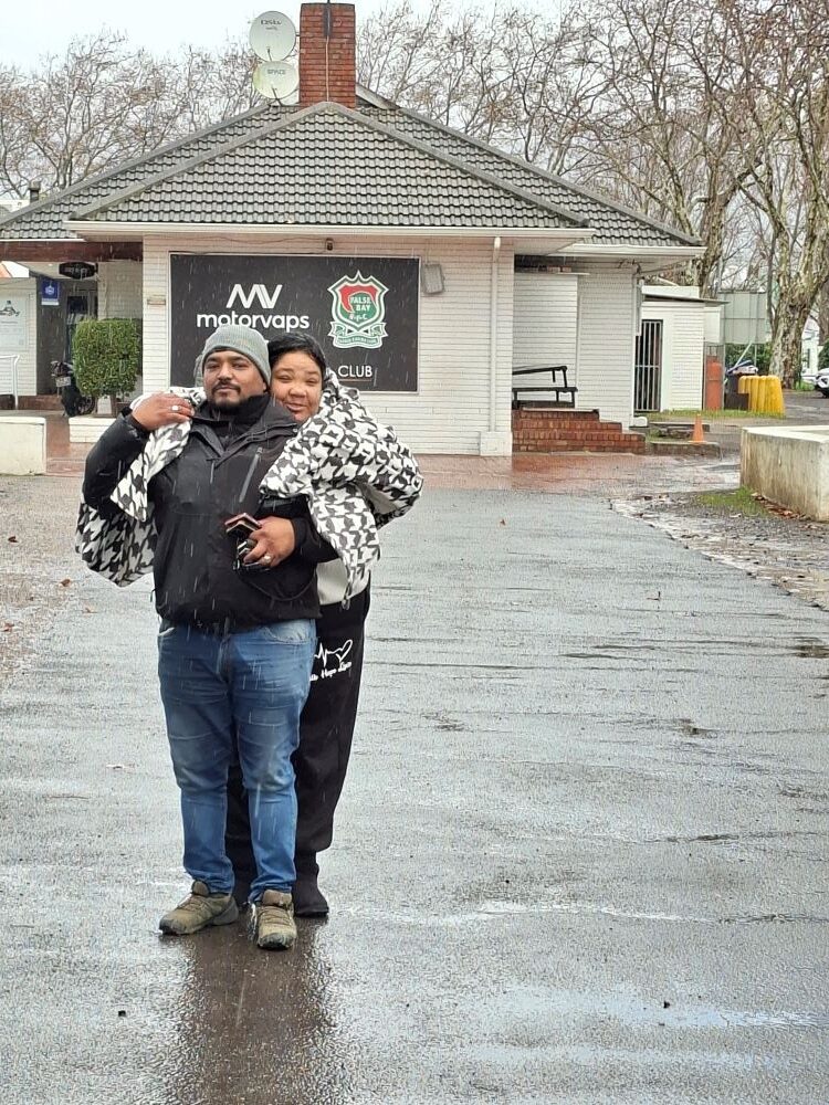 A portrait of Nolan Swanepoel and his wife hugging to stay warm on a wet asphalt driveway in front of the False Bay Rugby Club building during a rainy day out.