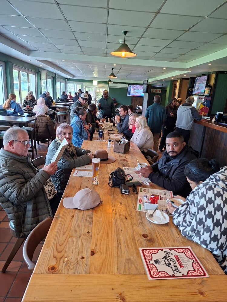 A long wooden table inside a bustling pub filled with classic car club members reading menus and ordering food after a rainy road trip.