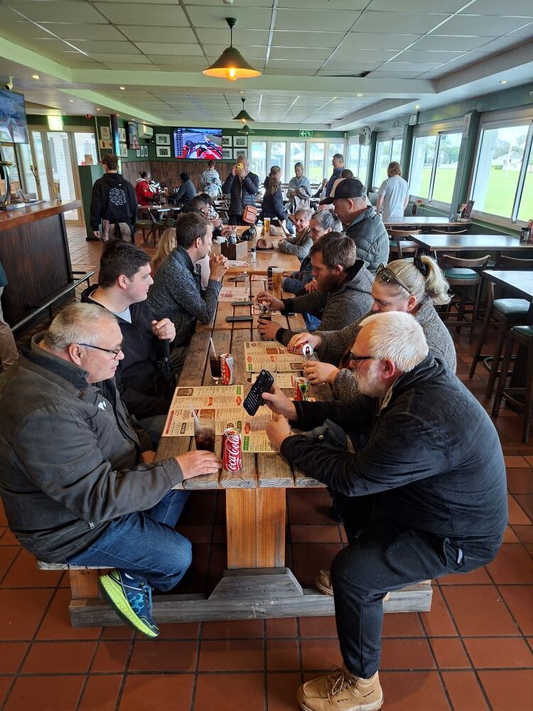 A high-angle shot of classic car club members sitting at a long wooden table inside a pub, talking, sharing drinks, and looking at a smartphone together.