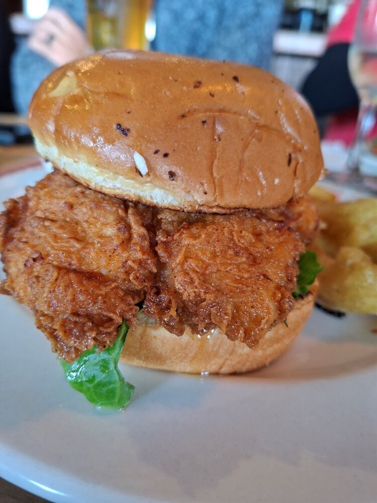 A close-up shot of a large, crispy golden fried chicken burger on a bun with lettuce, served with chips on a plate at a restaurant.