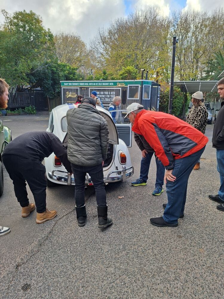 A group of classic car club members leaning in to inspect an open rear engine bay of a white classic VW Beetle with racing stripes, while the owner showcases the air-cooled engine setup at an outdoor parking lot meeting point.