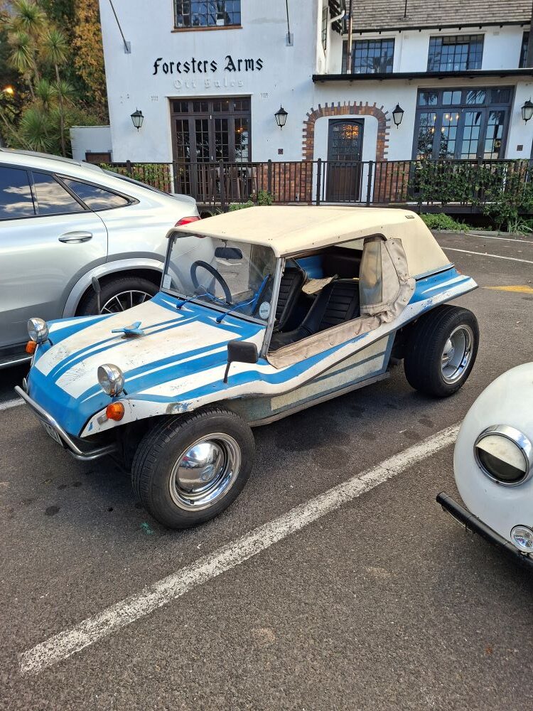 A classic white and blue Beamish beach buggy with a cream-colored soft top canopy, chrome wheels, and custom blue stripes, parked in front of the Foresters Arms building at a car club meeting spot.