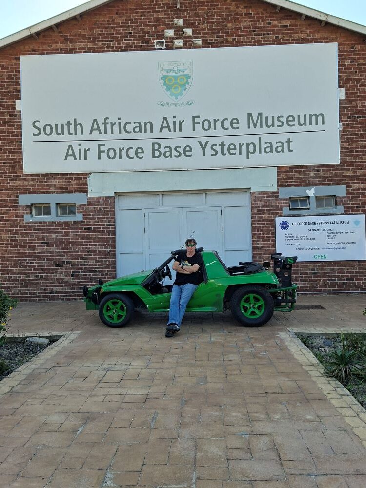 Anton Kleyn leaning against Shrek, a green Kango beach buggy with a water-cooled Nissan motor, parked outside the brick entrance of the South African Air Force Museum at Air Force Base Ysterplaat during a prerun for the Wild Buggers Beach Buggy Club.
