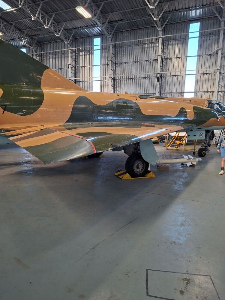 A vintage Atlas Cheetah fighter jet displaying a camouflage paint scheme inside a hangar at the South African Air Force Museum during a classic car run.